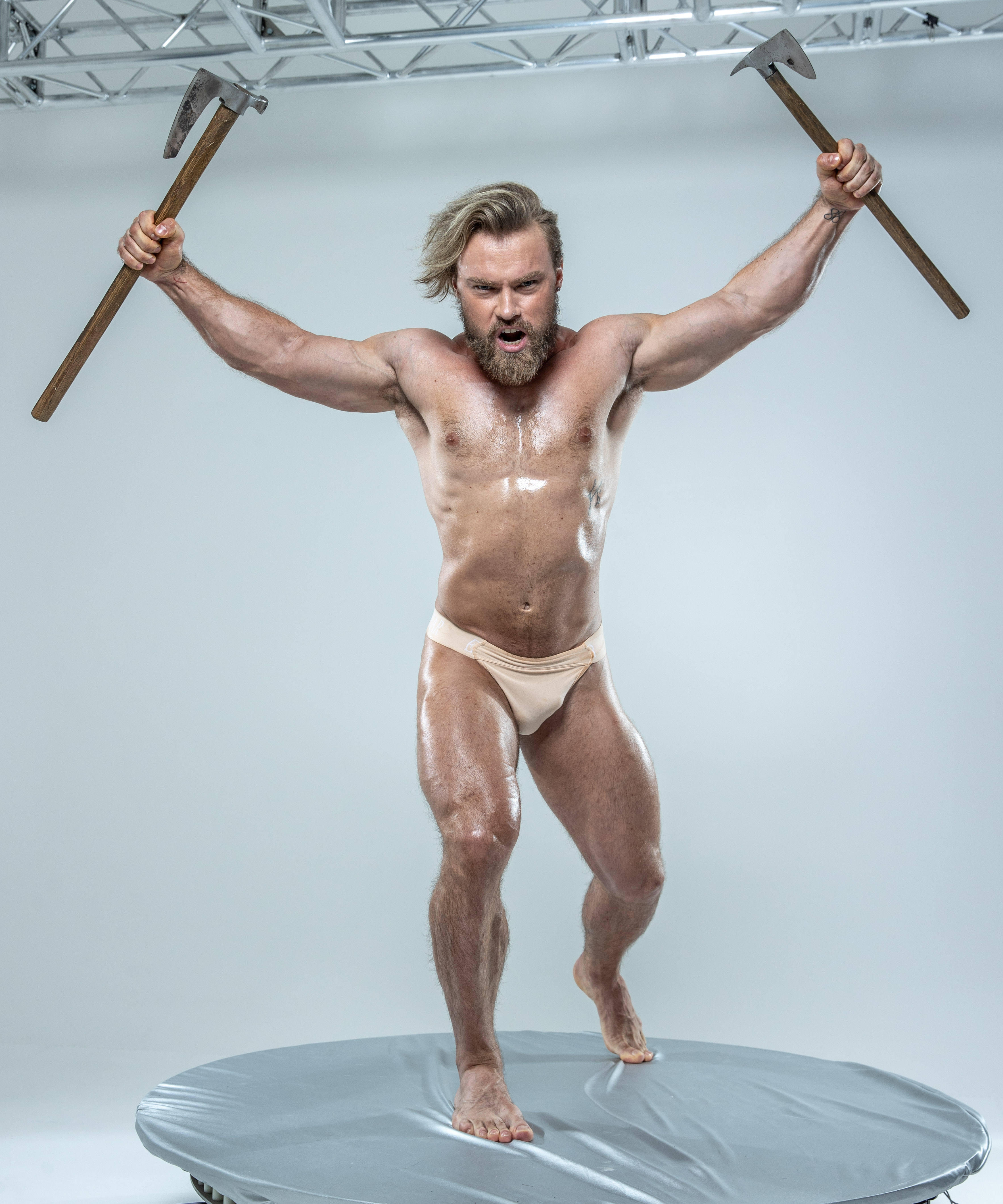 a muscular man with long blond hair and a beard is posing on a circular platform against a gray background. he is shirtless and wearing only beige underwear, his body glistening with sweat or oil. he holds an axe in each hand, raised above his head in a gesture of triumph or aggression. his mouth is open in a yell, and his muscles are tensed as if he is about to strike.  the lighting is bright and even, highlighting his physique and the axes.