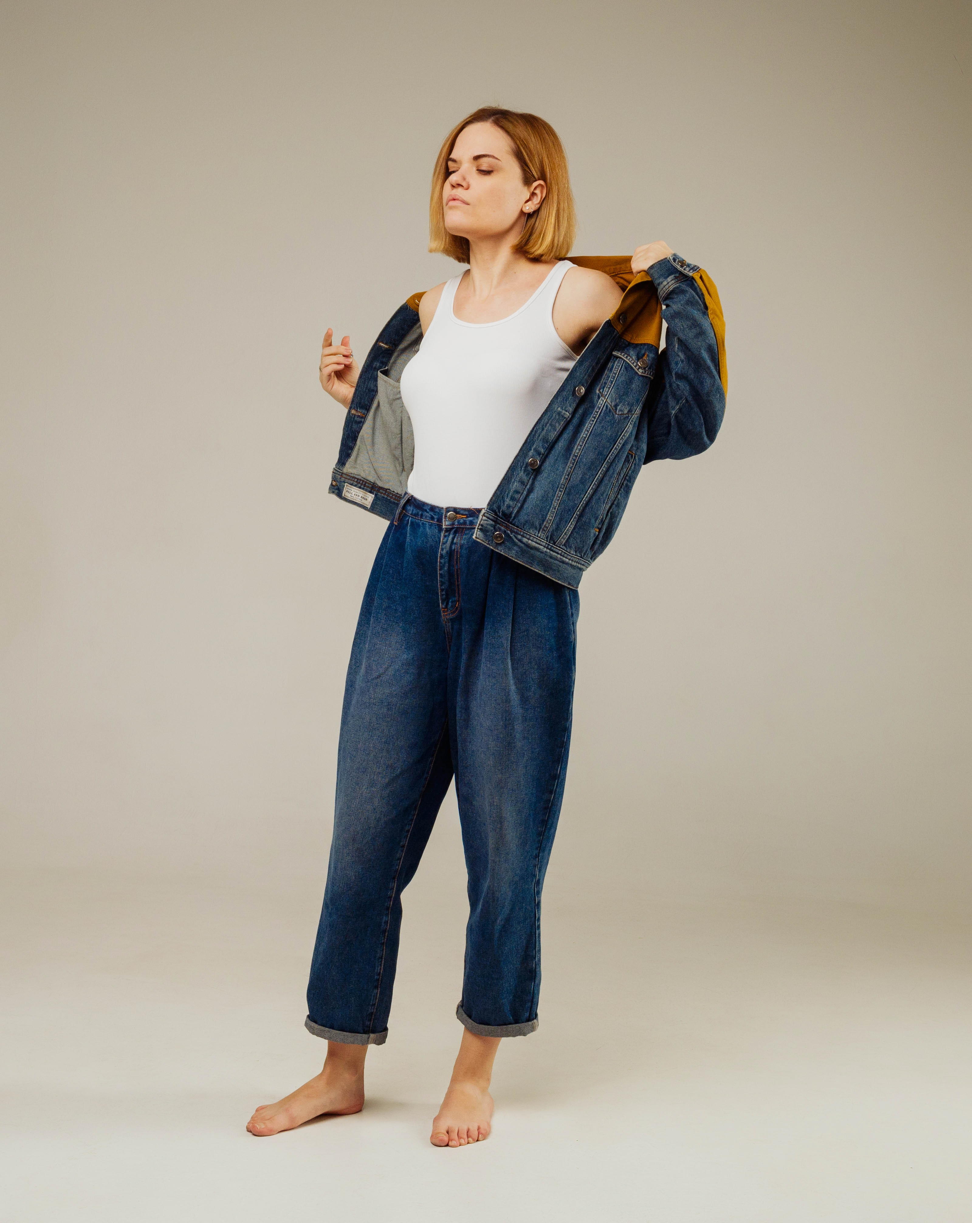 a woman with short blonde hair stands against a light gray background. she is wearing a white tank top, blue denim jeans and an oversized denim jacket. she is in the process of taking the jacket off, holding it by the collar with her hands. the jacket has a mustardcolored lining. the woman is barefoot, creating a relaxed and casual vibe. the image has a minimalist aesthetic, with the focus on the woman and her clothing.