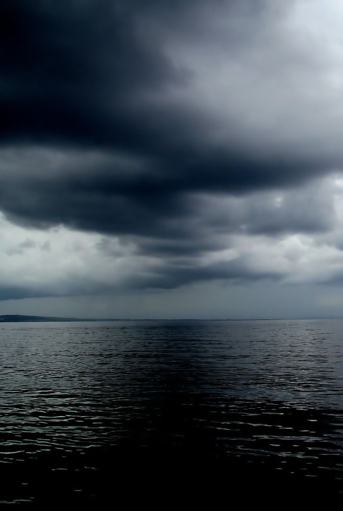 a dramatic, moody image of a stormy sky over a vast, calm ocean. the dark clouds fill the upper part of the frame, creating a sense of foreboding. the water below is still and reflective, mirroring the dark sky above. the image is a study in contrasts, with the dark sky and water offset by the faint glow of the horizon.
