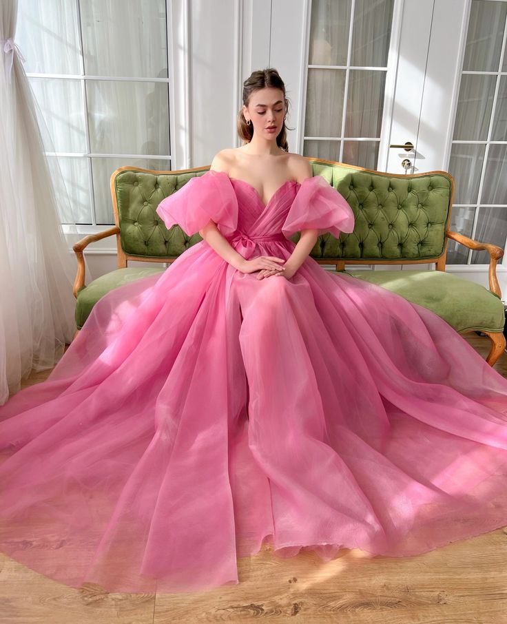 a young woman in a beautiful pink tulle gown sits on a green couch, she is posing for a photo. the dress is off the shoulder and has a  sweetheart neckline,  the bodice is fitted and the skirt is  full and flowing. her hair is pulled back and she has a natural makeup look.  the background is a white wall with a large window.
