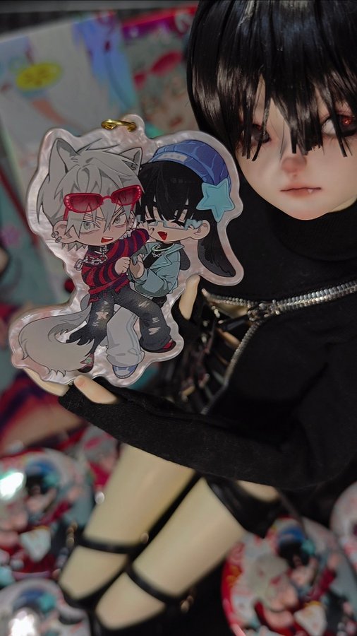 a closeup shot of a doll holding a charm of two anime characters. the charm is in a white circular frame with the two characters drawn in a chibistyle. the doll has long black hair and wears a black turtleneck sweater. the background is blurry, but you can see some other animerelated items.