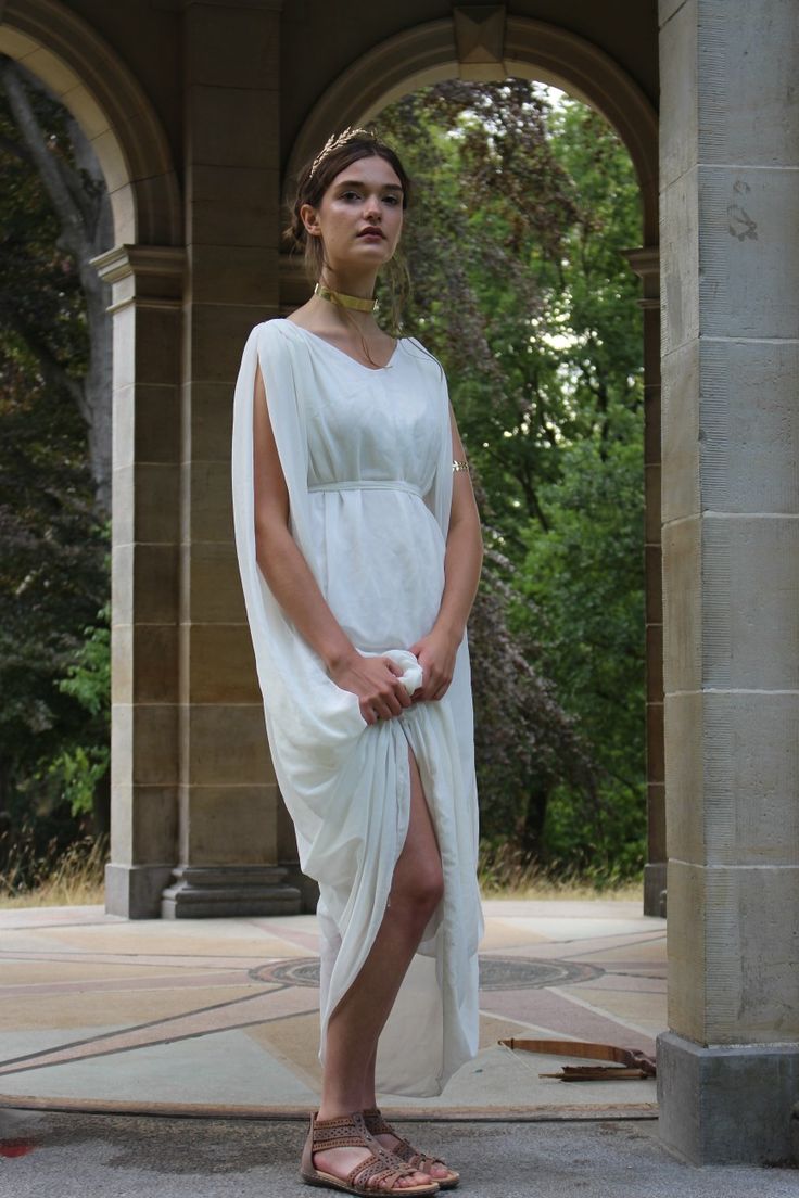 a young woman in a white ancient greekstyle dress stands between two stone columns in a park. she is wearing sandals, a gold headband, and a gold necklace. she is looking directly at the camera. the background is a lush green park.