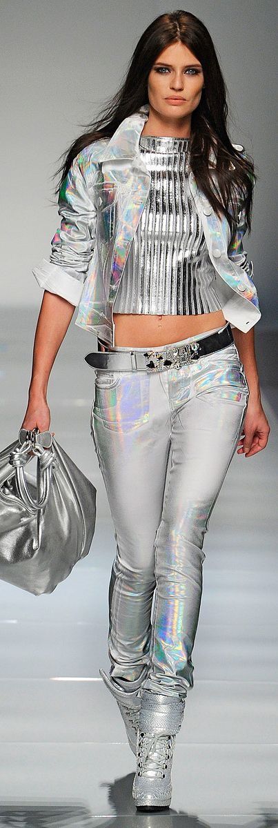 a model walks the runway in a silver jacket and jeans with matching boots. she has long dark hair and is carrying a silver handbag. she looks at the camera with a serious expression.  the background is a simple grey wall.