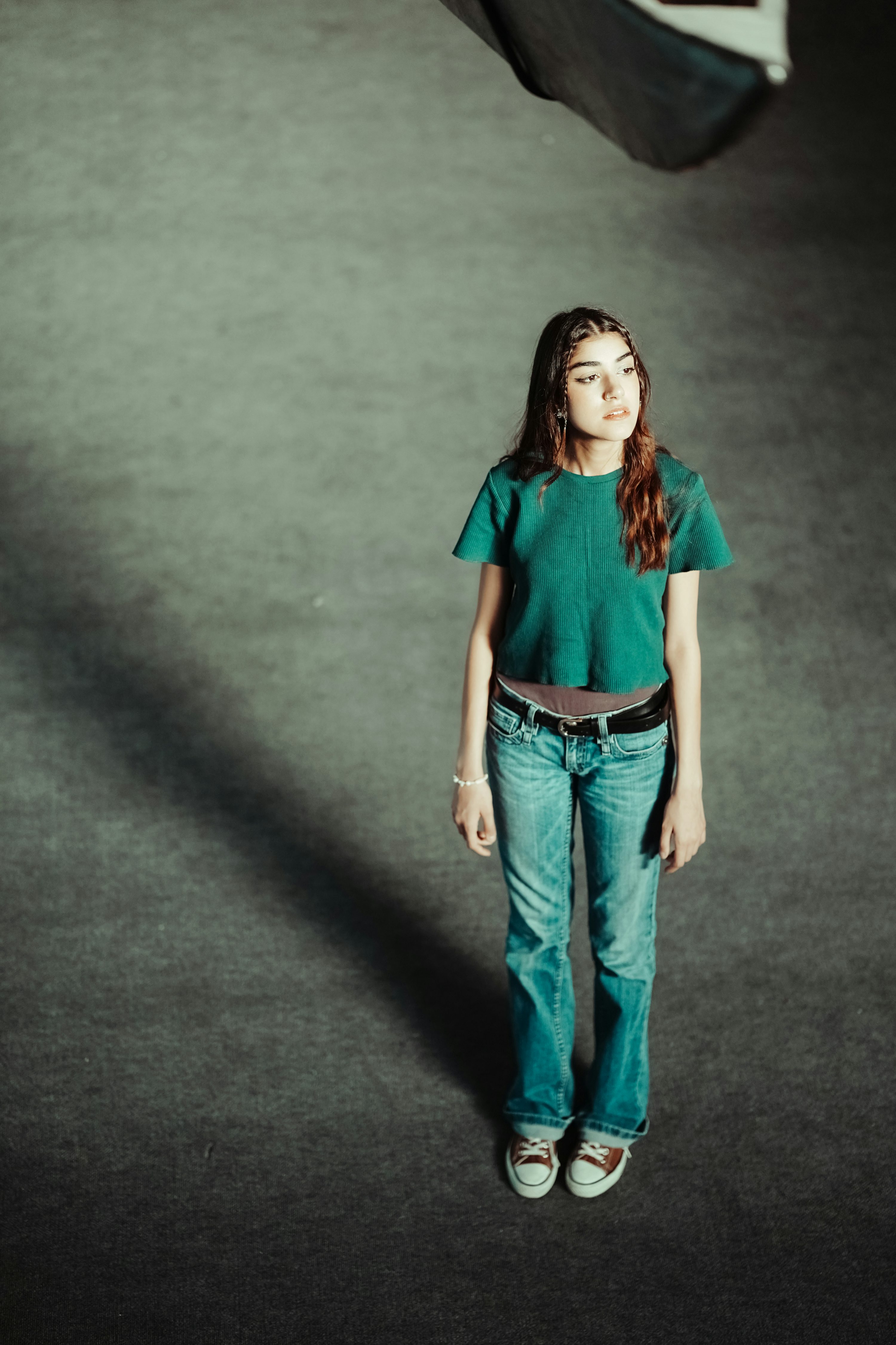 a woman stands in a large grey space, wearing a green tshirt, jeans, a belt, and sneakers. she is looking away from the camera. there is a shadow behind her and an object above her out of frame. the photo is taken from a high angle.