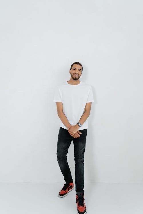 a fulllength shot of a smiling man wearing a white tshirt, dark jeans, and red and black sneakers. he has a neatly trimmed beard and short black hair. he is standing against a plain white background with his hands clasped. a watch is visible on his left wrist.