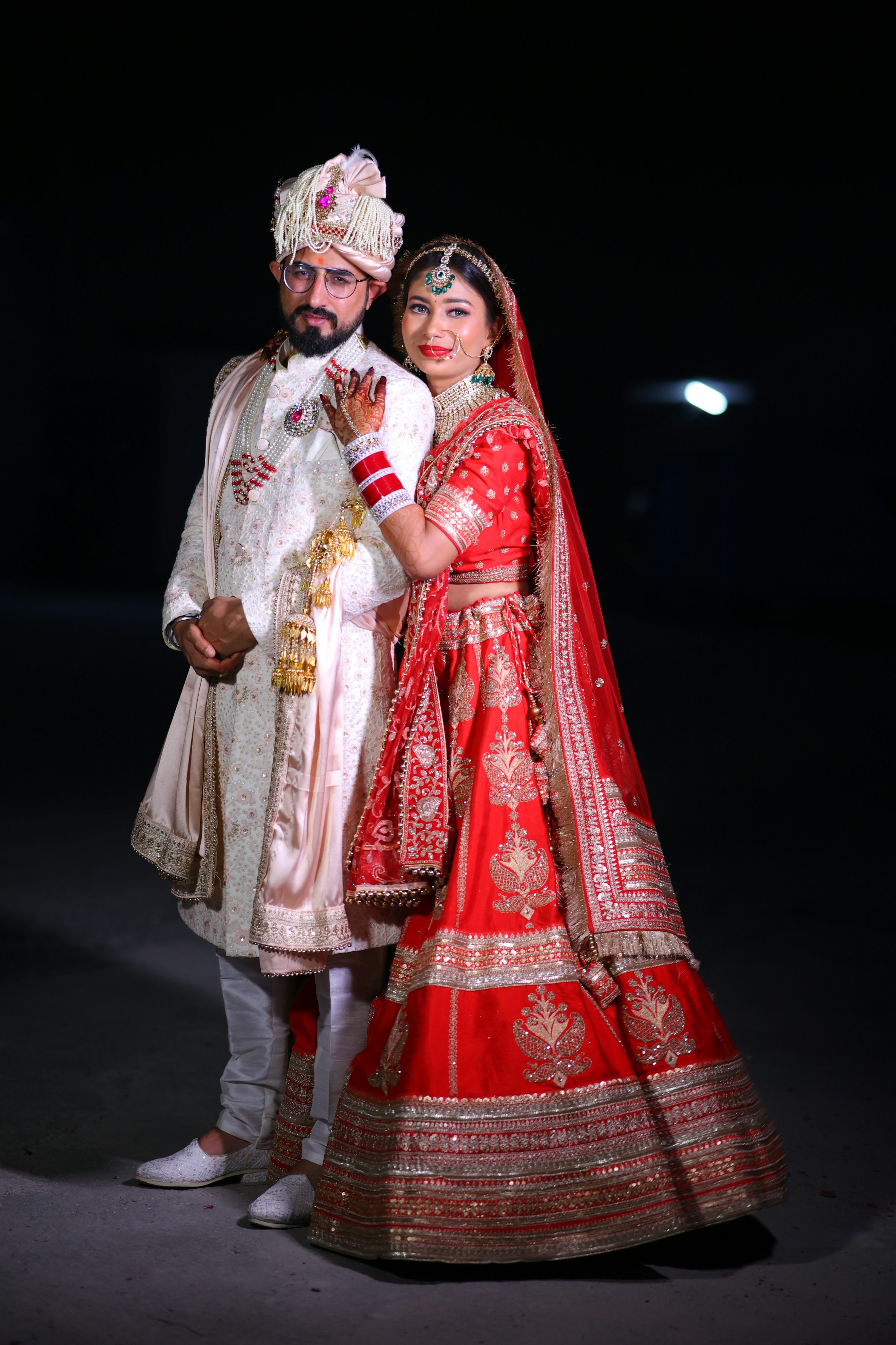 a stunning portrait of a newlywed indian couple. the bride is resplendent in a vibrant red lehenga, richly embellished with gold embroidery and intricate details.  her jewelry complements the attire perfectly. the groom is dapper in a creamcolored sherwani, a traditional long coat, paired with a matching turban (pagri). they stand close together, creating a picture of intimacy and happiness. the dark background highlights the couple's attire and adds a dramatic effect to the photograph. the image exudes a sense of elegance and traditional indian wedding celebrations.