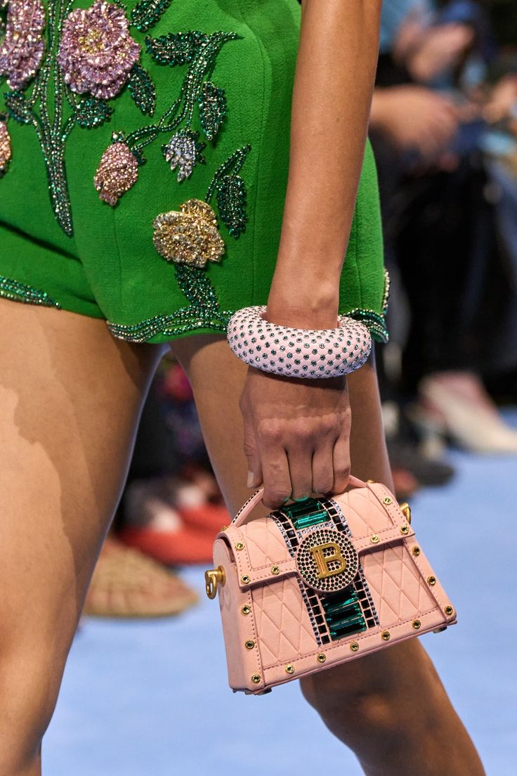 closeup view of a fashion model showcasing a pink balmain handbag.  the model is wearing a vibrant green, beaded shorts outfit. the handbag is a quilted design with a distinctive jewelencrusted detail on the flap. the model's hand is visible, adorned with a large pink beaded bracelet. the background is blurred, suggesting a fashion show setting. the overall image focuses on the details of the clothing and accessories, highlighting the luxury and embellishment.
