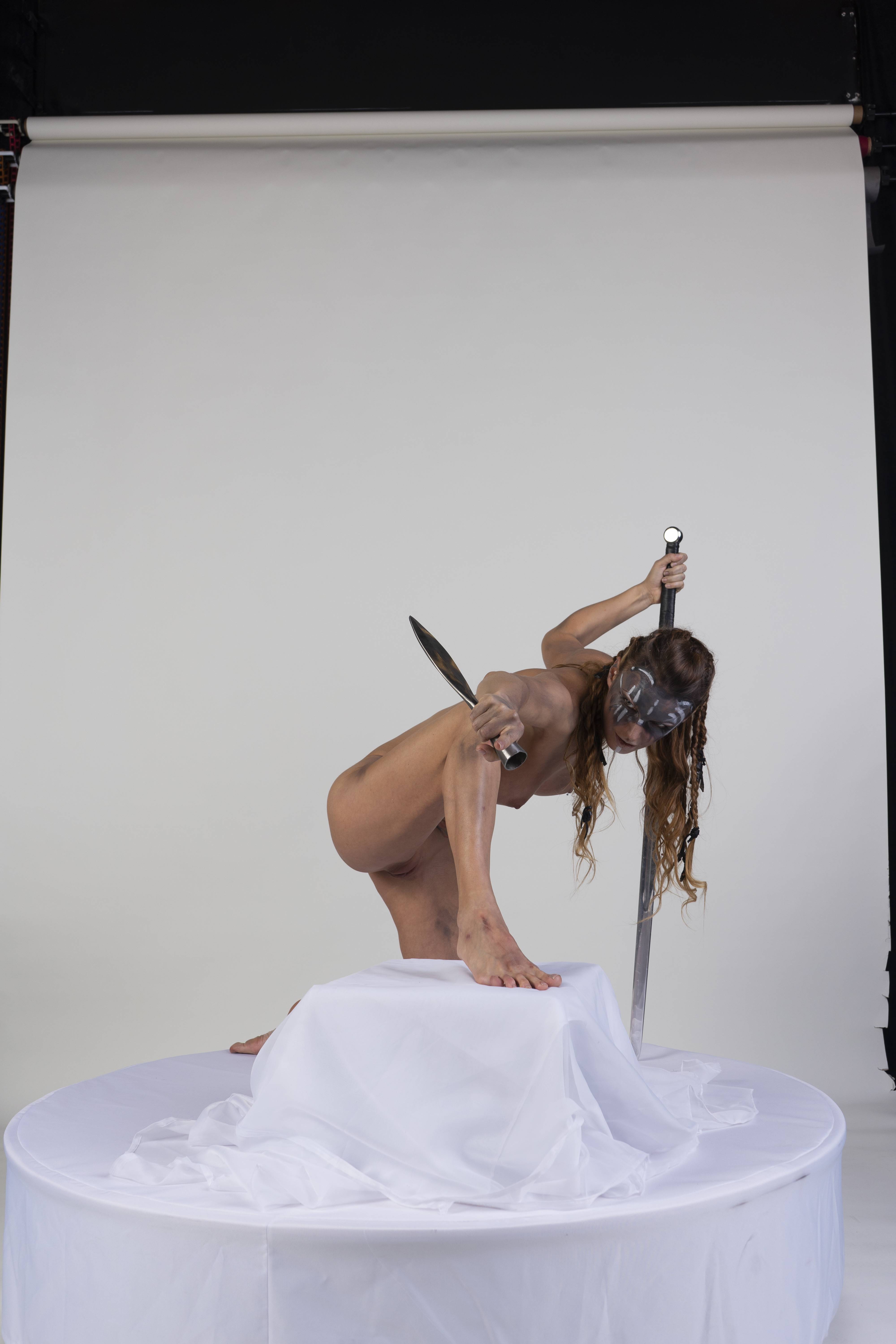 a nude female warrior with face paint is kneeling on a white draped platform in a studio setting. she is holding a sword in one hand and a knife in the other. the background is a plain white backdrop.