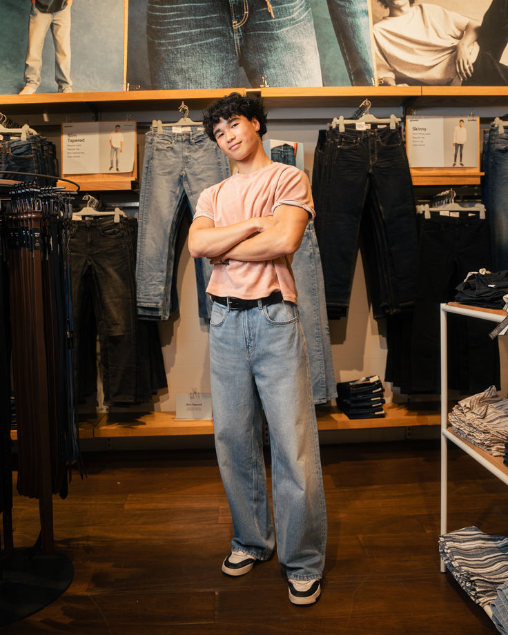 a young man is standing in a clothing store surrounded by racks of jeans. he's wearing a pink tshirt and light blue jeans, and looking at the camera. he's standing in front of a rack of jeans, showing off a variety of different styles and colors. he's casually dressed in a trendy and modern outfit. he's probably shopping for new clothes, or maybe he works there.