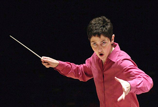 a female conductor in a pink shirt leads an orchestra with passion and intensity. she stands in the center of the stage, holding her baton aloft, her expression focused. the dark background highlights her presence and emphasizes the drama of the performance. her expressive hands and serious expression convey the energy and emotion of the music.