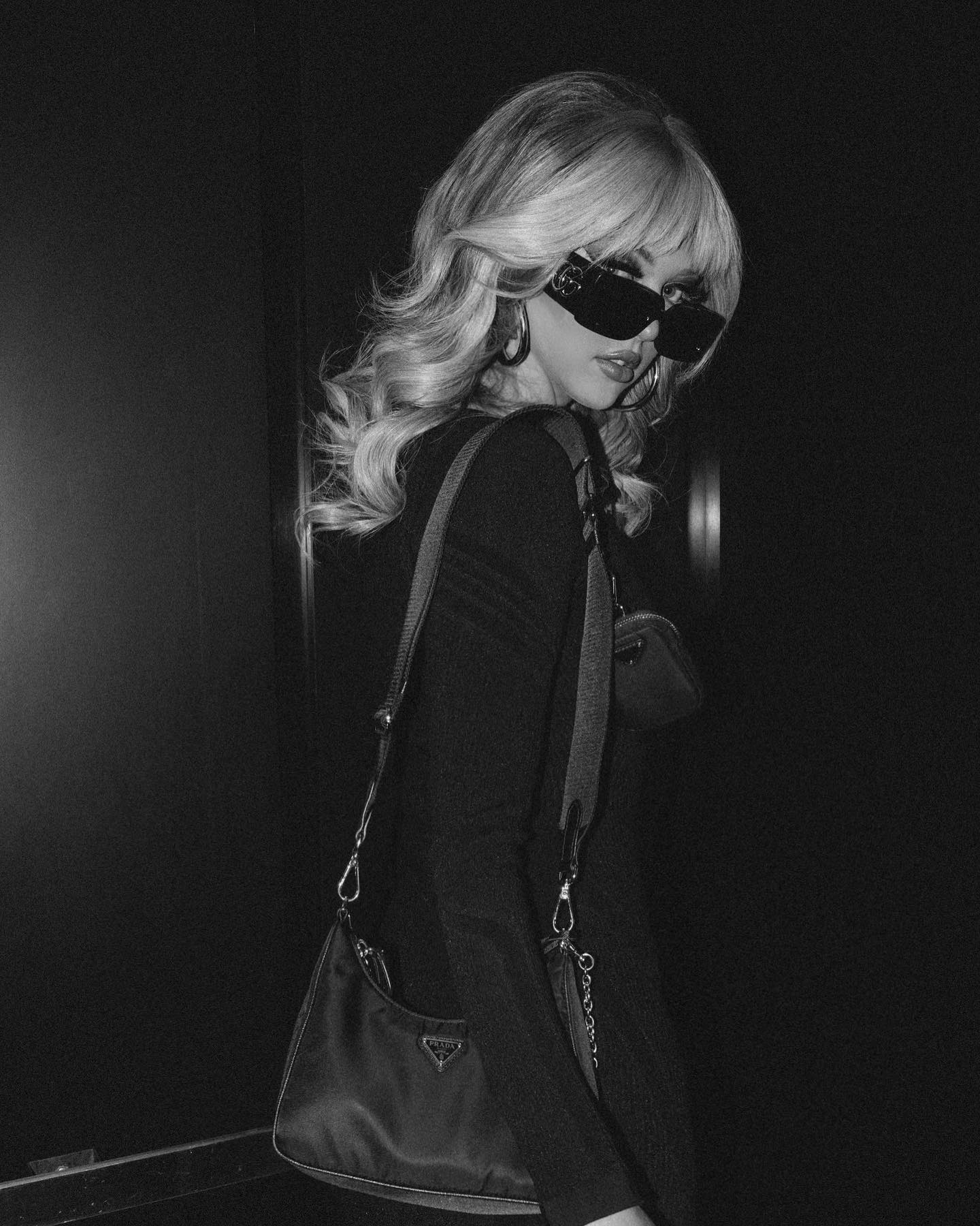 a black and white portrait of a stylish woman. she has blonde, wavy hair with bangs, and is wearing sunglasses and large hoop earrings. a black prada handbag is slung over her shoulder. she is wearing a dark, longsleeved top. the background is a dark, neutral tone.
