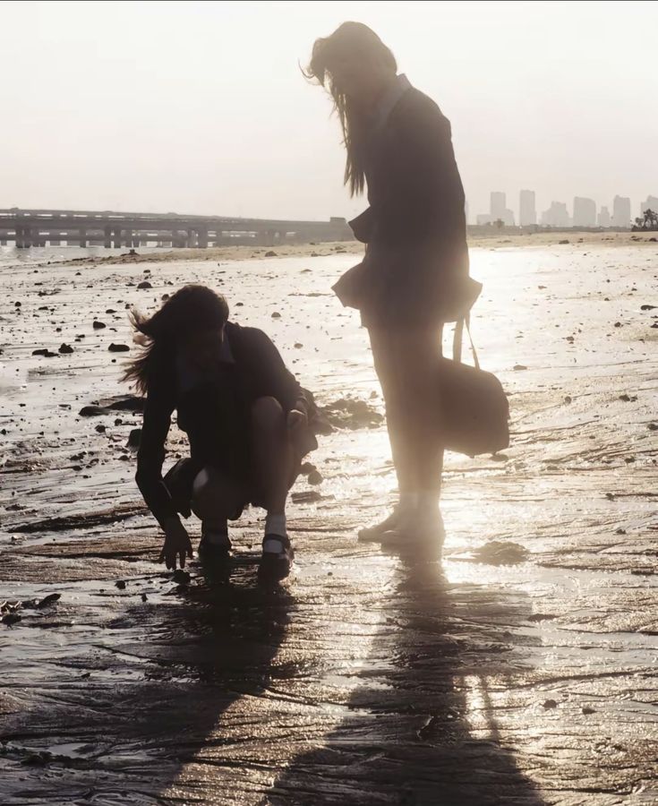 the image shows two girls, seemingly in school uniforms, silhouetted against a setting sun on a beach.  the beach appears to be a mudflat or intertidal zone, with wet sand showing textures and patterns.  a distant city skyline is visible in the background, suggesting an urban coastal area. the girls are engaged in some activity close to the ground, one crouching down while the other stands. the overall mood is peaceful and contemplative, enhanced by the backlighting and the soft light of the setting sun.  the shadows of the girls are prominently cast on the wet sand.  the scene is evocative of a tranquil moment of friendship and perhaps exploration.
