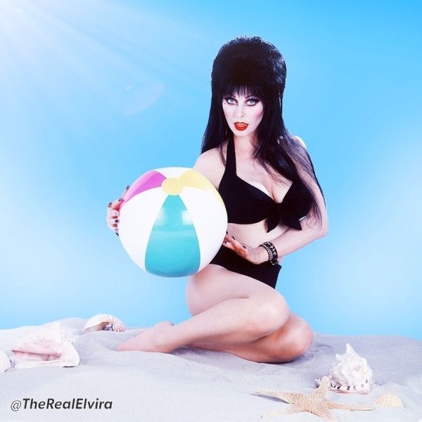 a photo of elvira, mistress of the dark, sitting on a beach in a black bikini, holding a beach ball. she is wearing red lipstick and has long, black hair. the photo is set against a blue sky with clouds. the image evokes a sense of fun and summer vacation.