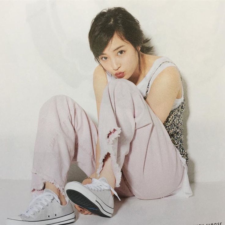 a young asian woman with short black hair is sitting on the floor with her legs crossed. she is wearing a white sleeveless top, pink denim pants, and white converse sneakers. she is looking at the camera with a pouting expression. she is in a studio setting with a white background.