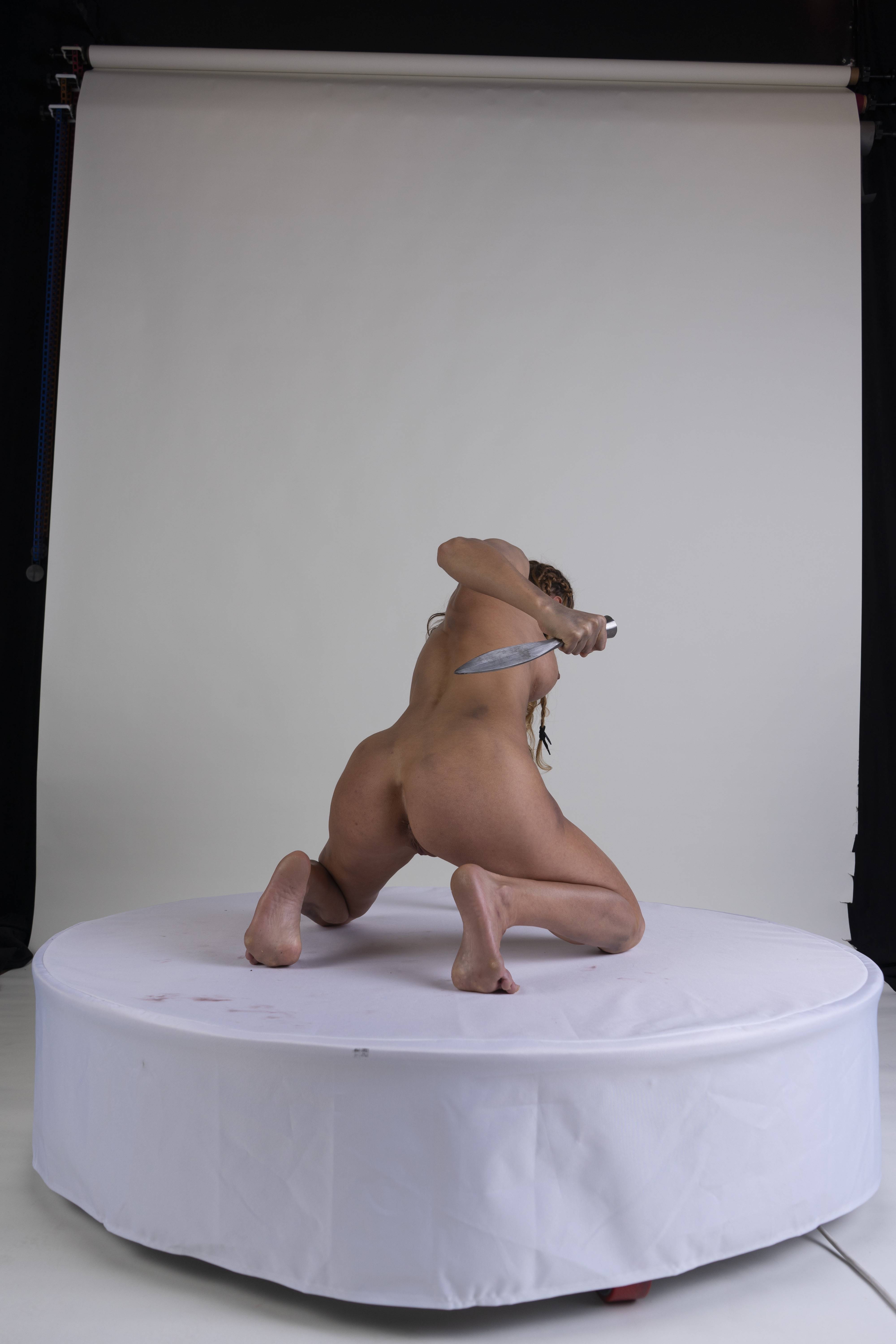 a nude woman is kneeling on a white circular platform indoors, holding a knife behind her back. the background is a plain white studio backdrop.