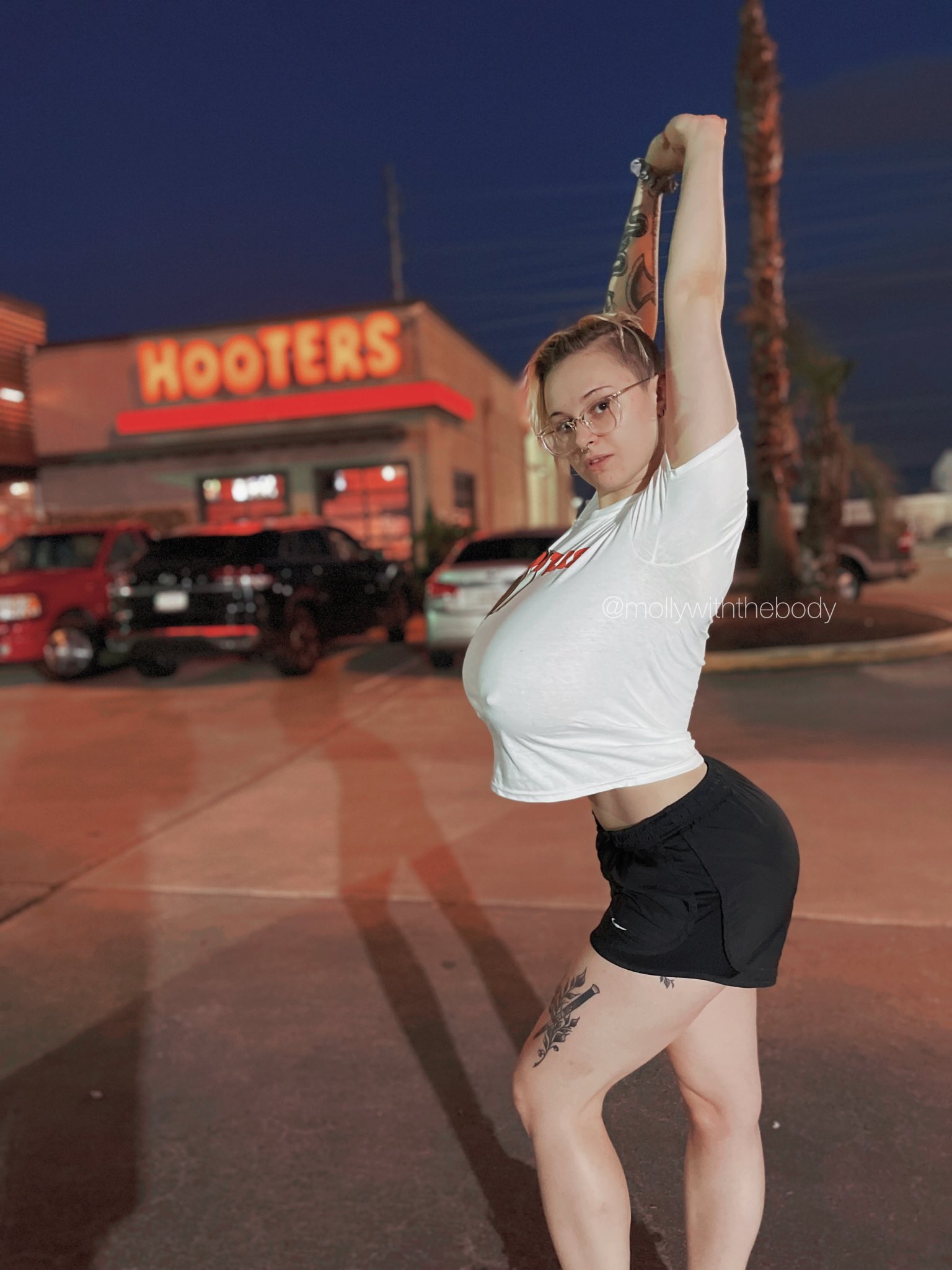 the image shows a young blonde woman wearing a white tshirt and black shorts, standing in a parking lot at night in front of a hooters restaurant.  she's stretching one arm overhead, showing a tattoo on her leg. her expression is confident, and she appears relaxed. the background is slightly blurry, focusing attention on the woman. the overall mood is casual and summery.