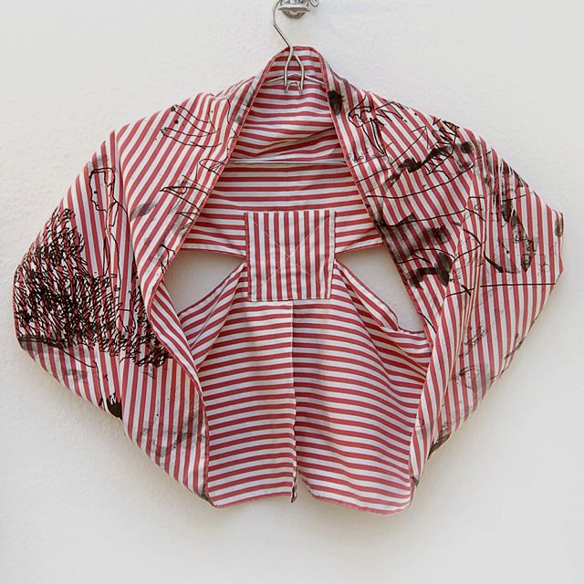 a red and white striped fabric garment hangs on a silver hanger against a white wall. the garment, resembling a kimono or jacket, features abstract black drawings and markings. the striped pattern is consistent throughout, and the garment appears to be handmade or customized with the unique artistic additions.