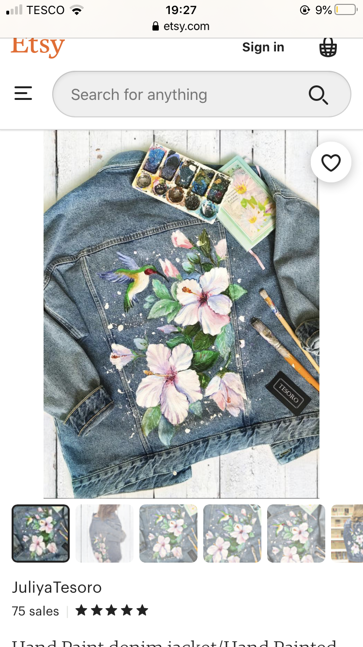 a denim jacket is displayed on a white background. it is painted with pink flowers, green leaves and a hummingbird. there are paint brushes and a watercolor set on top of the jacket. below the main image, there are six smaller images showing the jacket from different angles.