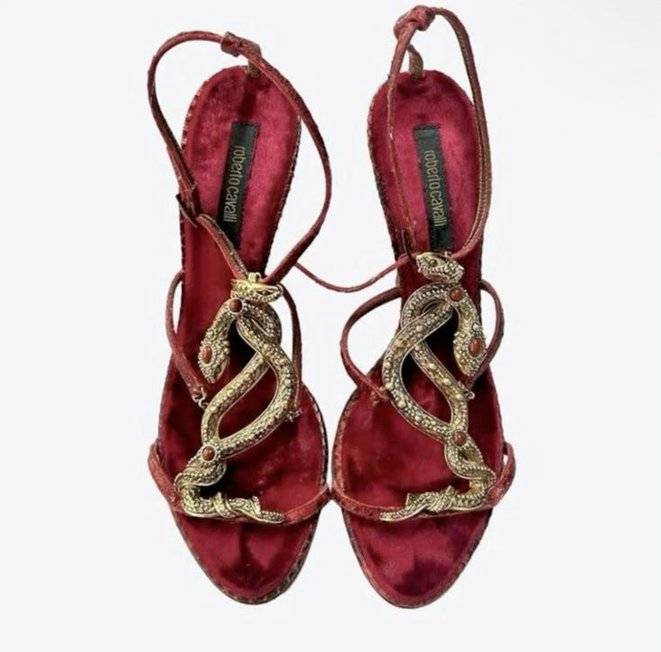 a pair of roberto cavalli women's high heel sandals are shown. the sandals are made of red velvet and feature gold snakeshaped embellishments with red gemstones. the shoes have ankle straps and are elegant in style. they are positioned to display both shoes, with the heels slightly tilted inward. the background is a simple white.