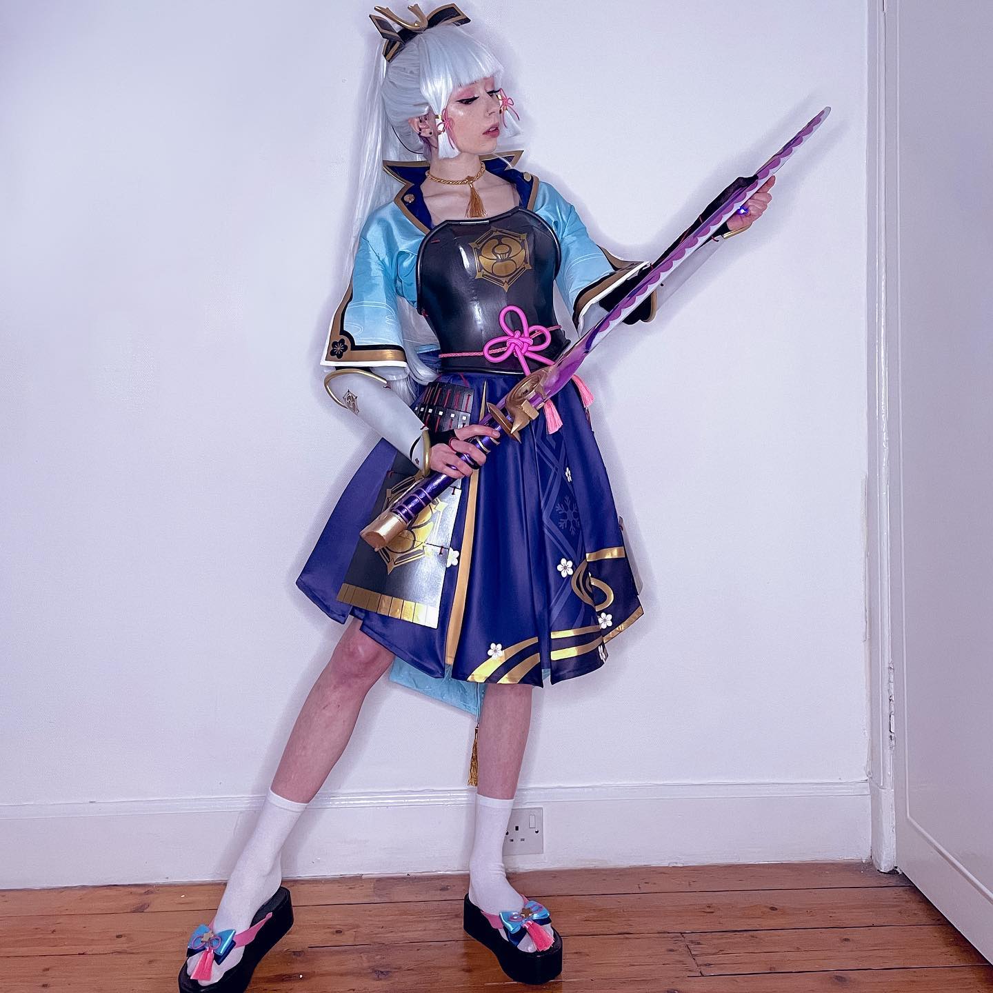 a fullbody shot of a young woman cosplaying as a female samurai character. she is wearing a detailed, vibrant costume that incorporates elements of japanese clothing and armor.  the costume features a blue and purple skirt, a metallic chest piece, and elaborate arm pieces. she holds a large, ornate purple and gold katana. her long, white hair is styled in a way that complements the character's aesthetic. she is wearing white socks and colorful geta sandals. the background is a simple white wall and wooden floor, which contrasts with the vibrant details of the costume and highlights the cosplay. the overall image is welllit and professionally presented, showcasing the detail and quality of the cosplay.