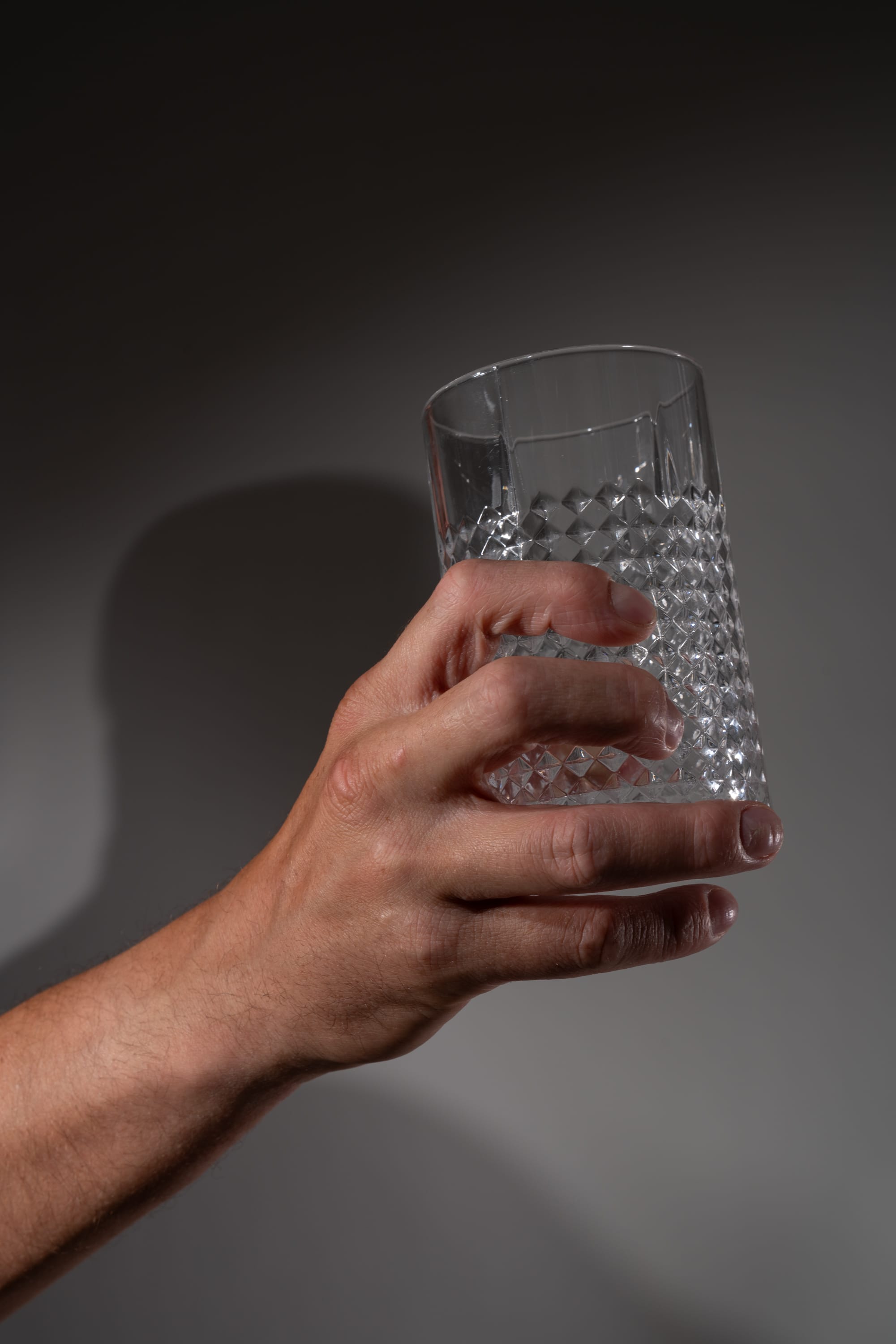 a hand holding a clear glass with a diamond pattern. the background is gray with a shadow cast by the hand and glass.