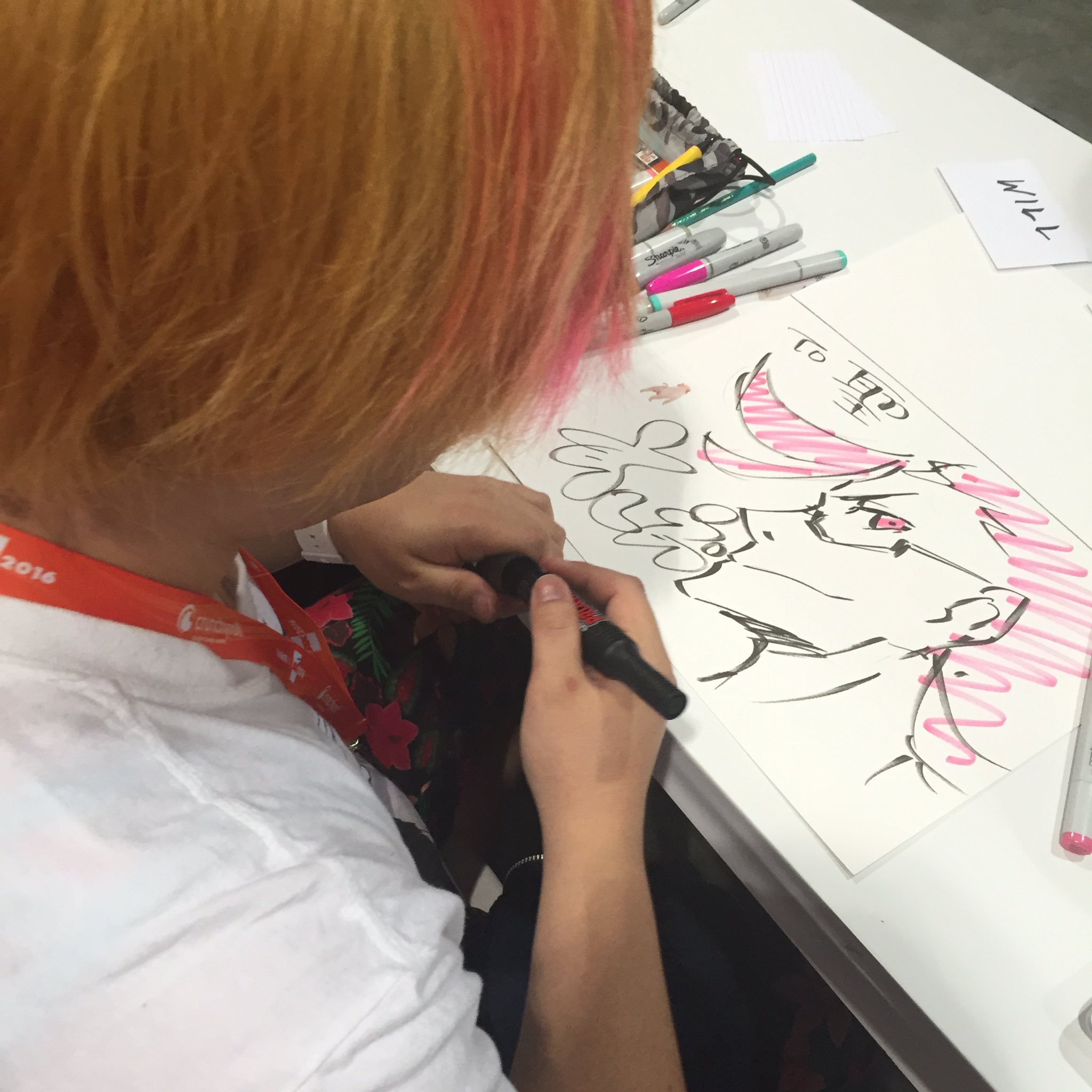 a person with dyed orange and pink hair is concentrating on drawing a furry character with pink and black markers. the drawing is on a white paper on a white table. there are some colorful markers scattered around the drawing and a name card that says "will". the environment appears to be bright and indoors, likely an artist alley at a convention.