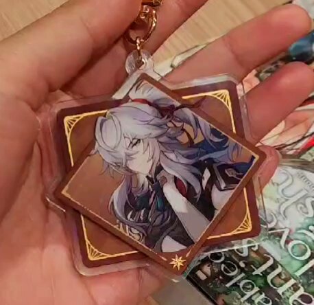 a closeup shot of an anime character keychain. the keychain is made of clear plastic with a gold border and features a character from the anime series. the character is depicted in a stylized, cartoonish fashion and is surrounded by a decorative border. the keychain is held in the hand of a person who appears to be a fan of the anime series. the keychain is a popular item among anime fans and can be used to decorate bags, keychains, or other items.