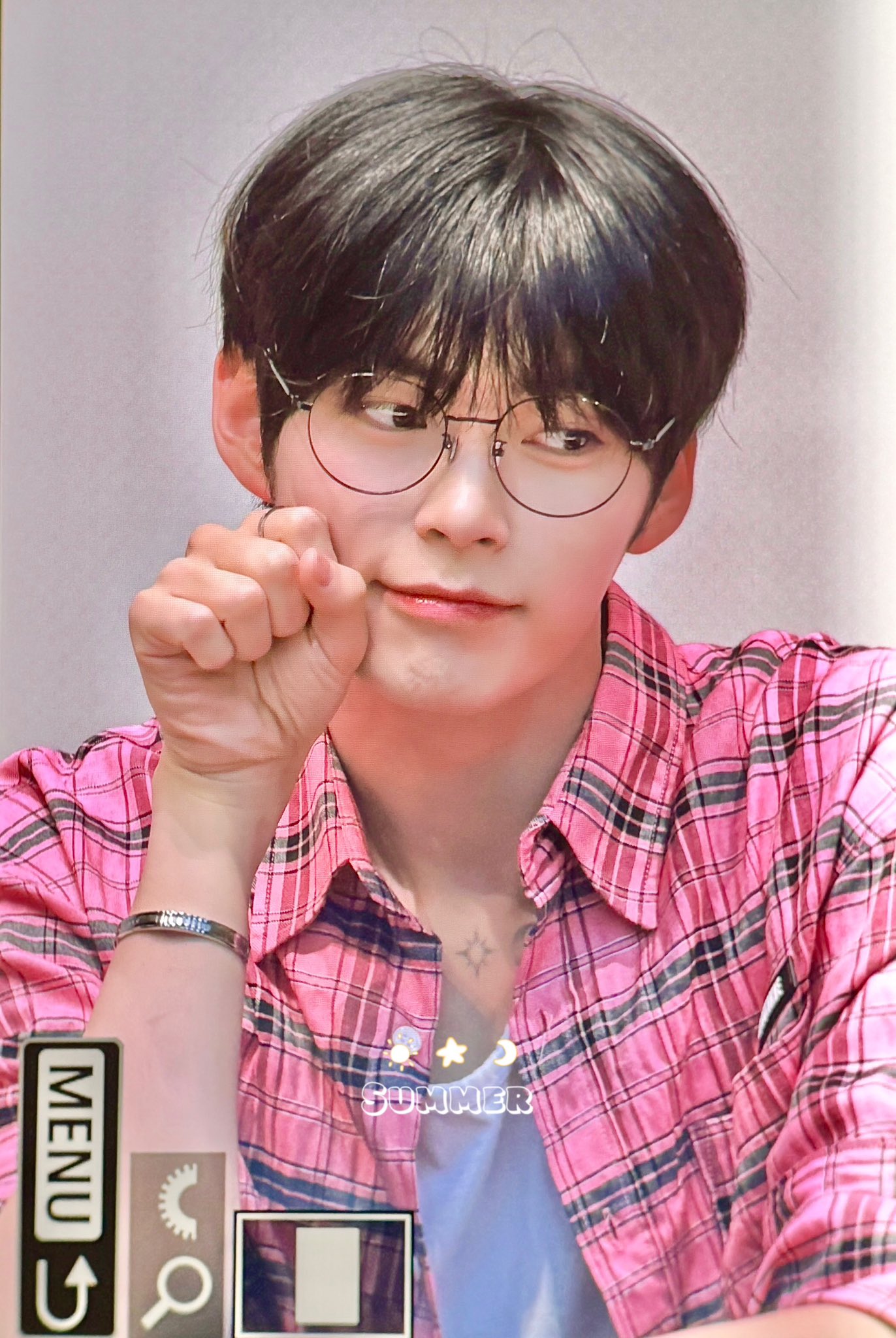 a selfie of a young man with black hair wearing glasses and a pink plaid shirt.  he has a silver bracelet on his right wrist and a small tattoo below his collarbone. he is posing with his left hand near his face. the background suggests he is indoors.