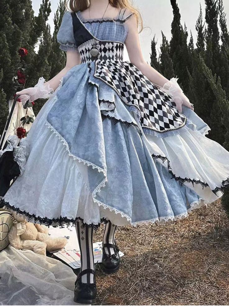 a young woman poses in an elaborate blue and white lolita fashion dress. the dress features a mix of patterned and solid fabrics, with a checkered pattern prominently featured on the skirt. the dress has a tiered, ruffled design with lace trim. she is standing on a blanket on the ground with a basket and a teddy bear nearby. the background is a natural setting with green foliage.  the lighting is soft and natural, creating a dreamy atmosphere.