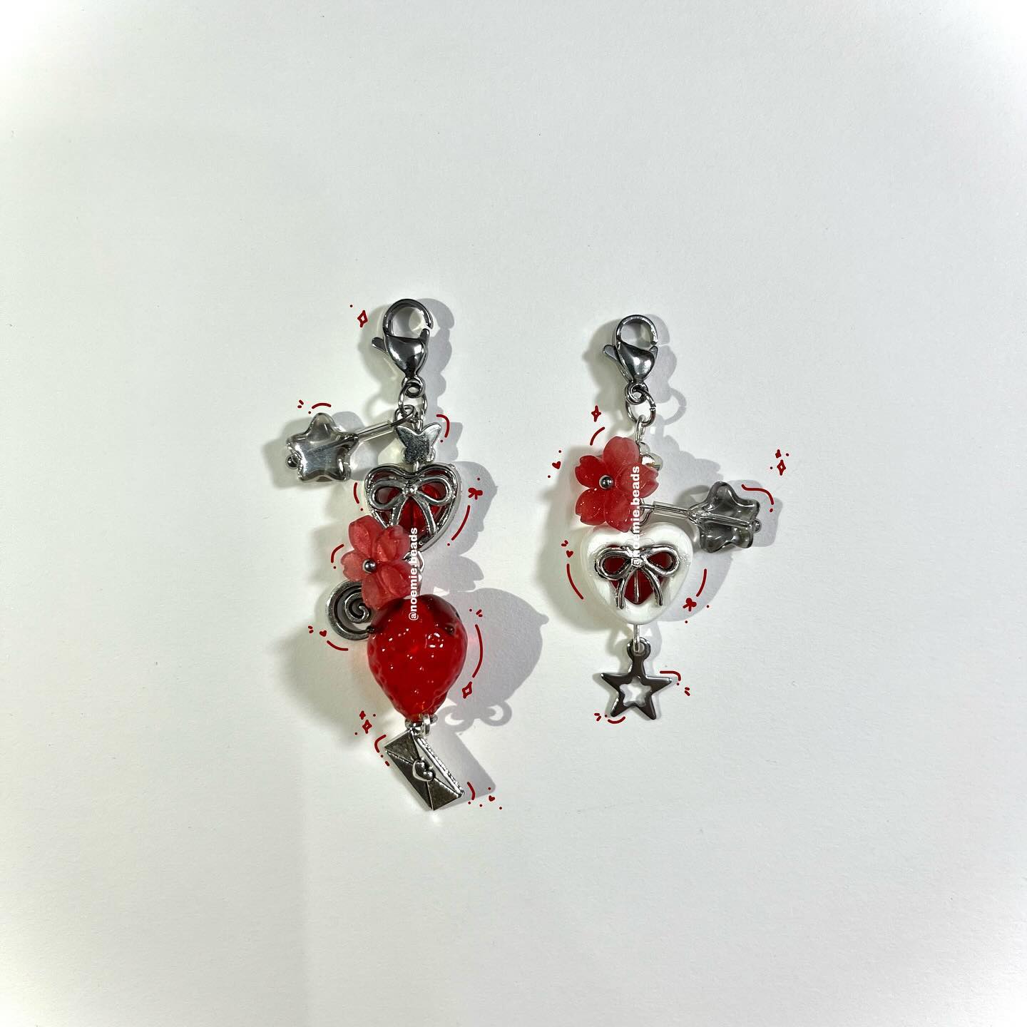 two handmade charm earrings are displayed on a white surface. the earring on the left features a silver clasp, a silver star charm, a heartshaped bow charm, a red flower, a red strawberry bead, and a silver letter charm. the earring on the right features a silver clasp, a red flower, a white heartshaped bead with a red bow, a silver key charm, and a silver star charm.