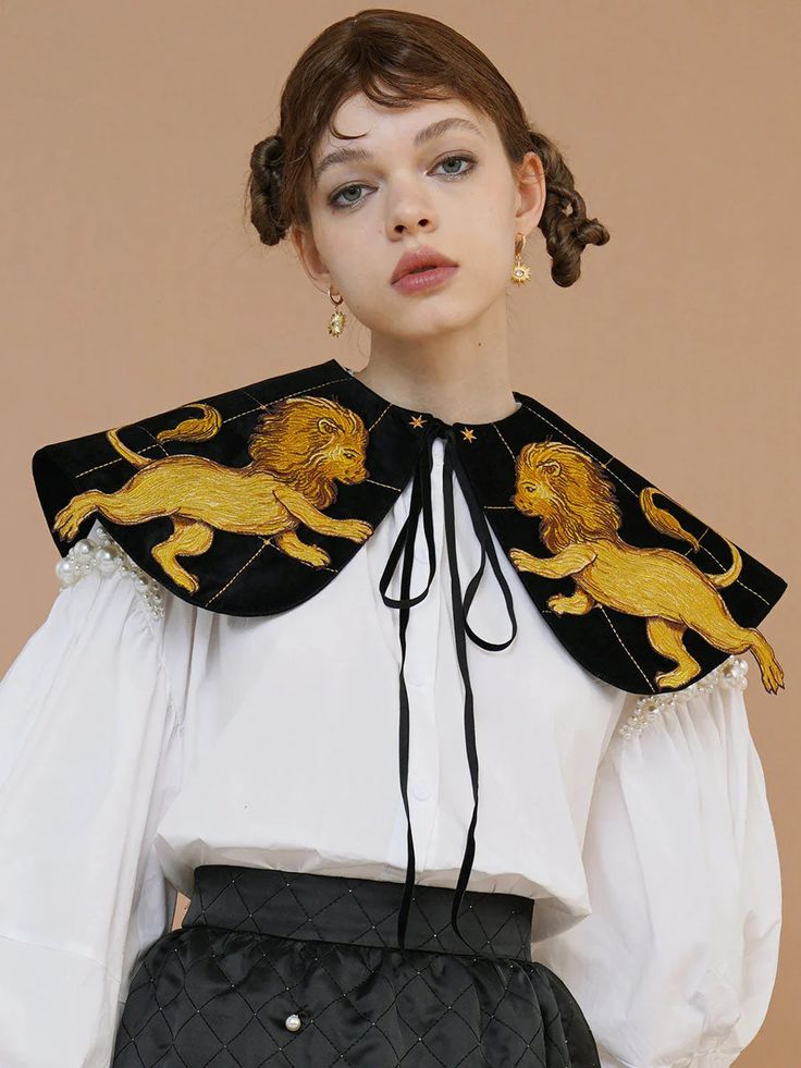 a young woman with a styled updo hairstyle is the focus of the image. she's wearing a white puffsleeved blouse, accessorized with a striking black velvet collar embellished with gold embroidered lions.  the collar has a tied bow at the front and small pearl details on the edges. she also wears a black quilted skirt and gold earrings. the backdrop is a neutral beige tone, highlighting the details of her clothing. the overall style is elegant and slightly vintageinspired.  the photo is a professional, likely for a fashion publication.