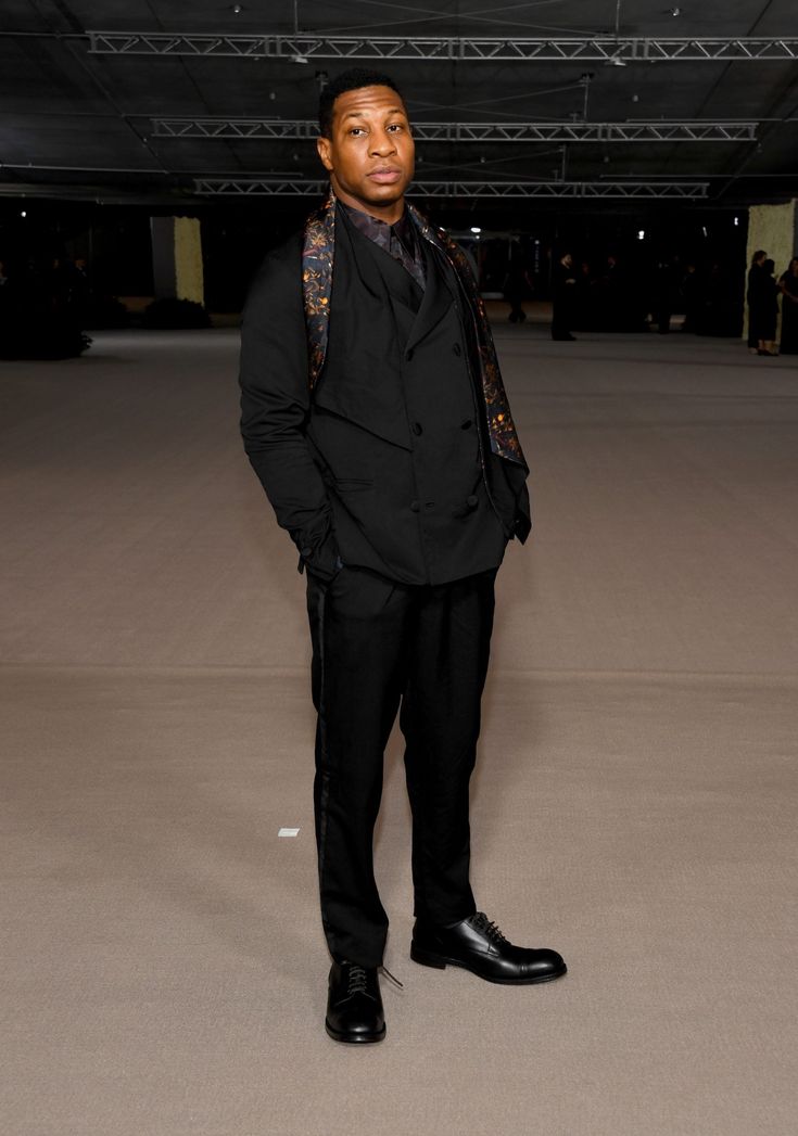 a fullbody shot of a man in a stylish black suit and black shoes, standing confidently in an indoor setting.  he's wearing a dark patterned scarf around his neck and his pose suggests he's at an event like a fashion show. the background is a large, empty space with hints of other people in the distance. the overall impression is one of sophistication and elegance.