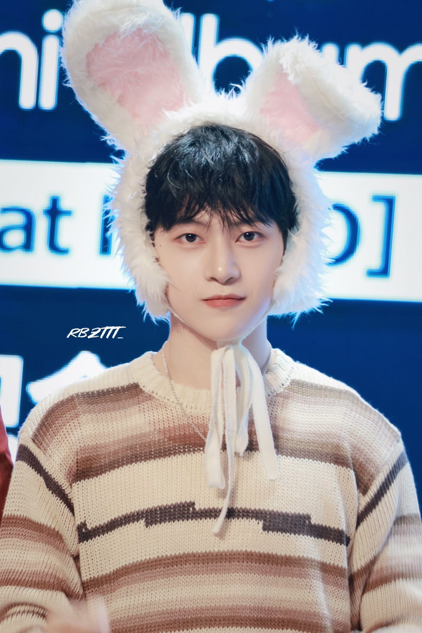 a closeup photo of an asian man wearing a fuzzy white bunny hat. he has short, dark hair and is wearing a beige and brown striped sweater. the background is blurred and there is text visible. the image appears to be taken at a fansign event.