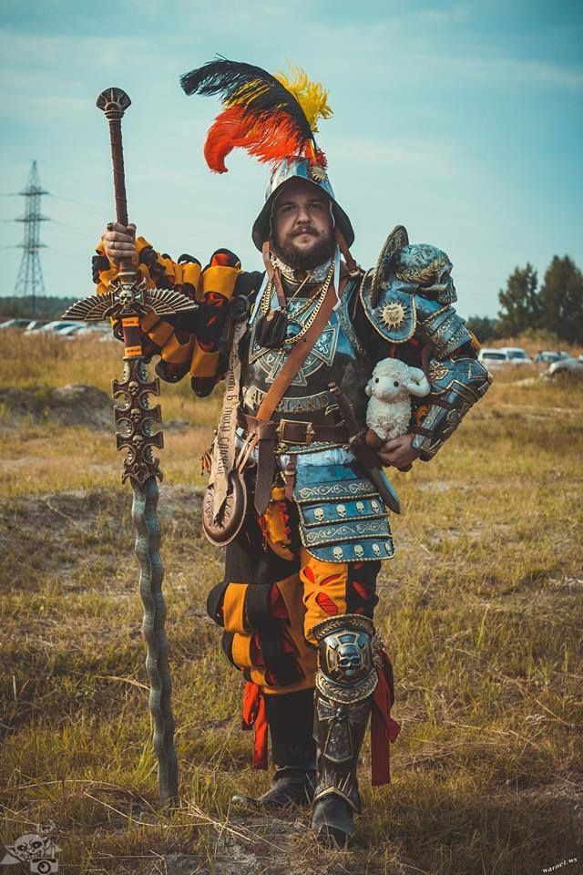 a man in a medieval knight costume, complete with armor, helmet, and a sword, is standing in a field. he is holding a plush lamb in his hand. he is looking at the camera with a serious expression. the costume is very detailed and intricate. the man is wearing a black, red, and yellow costume, with a lot of metal details. the costume is probably made of a thick fabric, and it looks very heavy. the man is standing in a field with green grass and some trees in the background. the photo is taken from a low angle, and the man is looking at the camera. the image is well lit, and the colors are vibrant. it's a great image for a cosplay photo shoot.