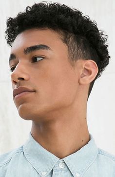a portrait of a young black man with short, curly hair, styled in a fade haircut. he has brown eyes, a defined jawline, and is wearing a light blue denim shirt. the lighting is soft and the background is white, giving the image a clean and minimalist aesthetic.