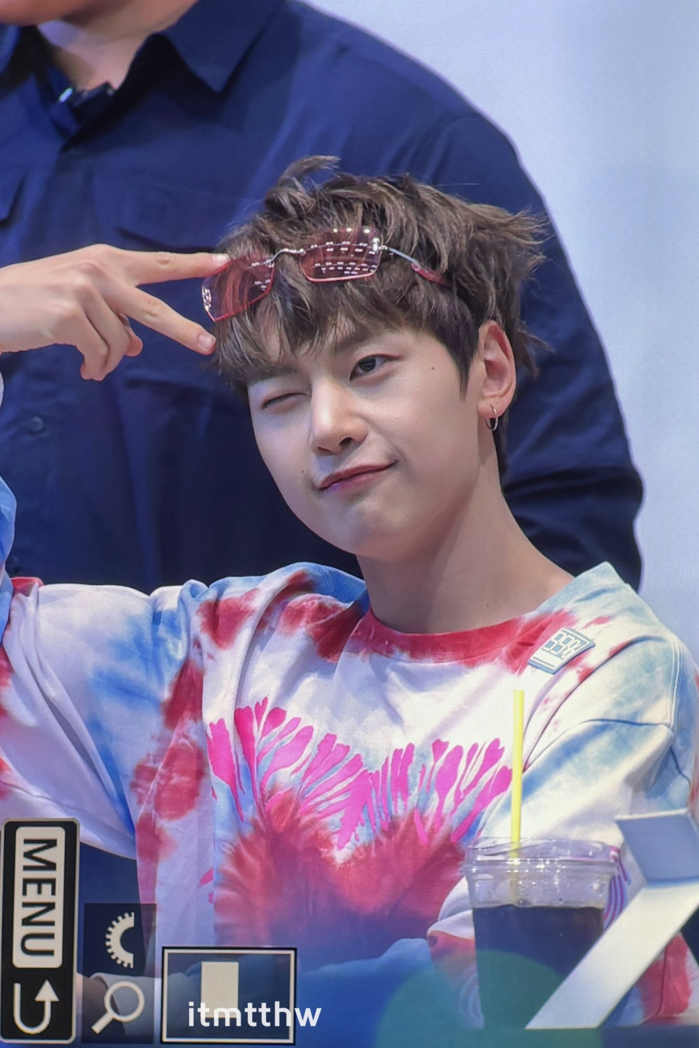 the image shows kim donghyun of the kpop group ab6ix at what appears to be a fansign event. he is wearing a tiedye shirt and red tinted sunglasses pushed up on his head. he is making a peace sign with his right hand and winking at the camera with a playful expression. donghyun has short brown hair styled up and is wearing a small earring in his left ear. in front of him is a partially visible plastic cup with a dark liquid, possibly iced coffee.  the setting suggests an indoor location with bright artificial lighting.