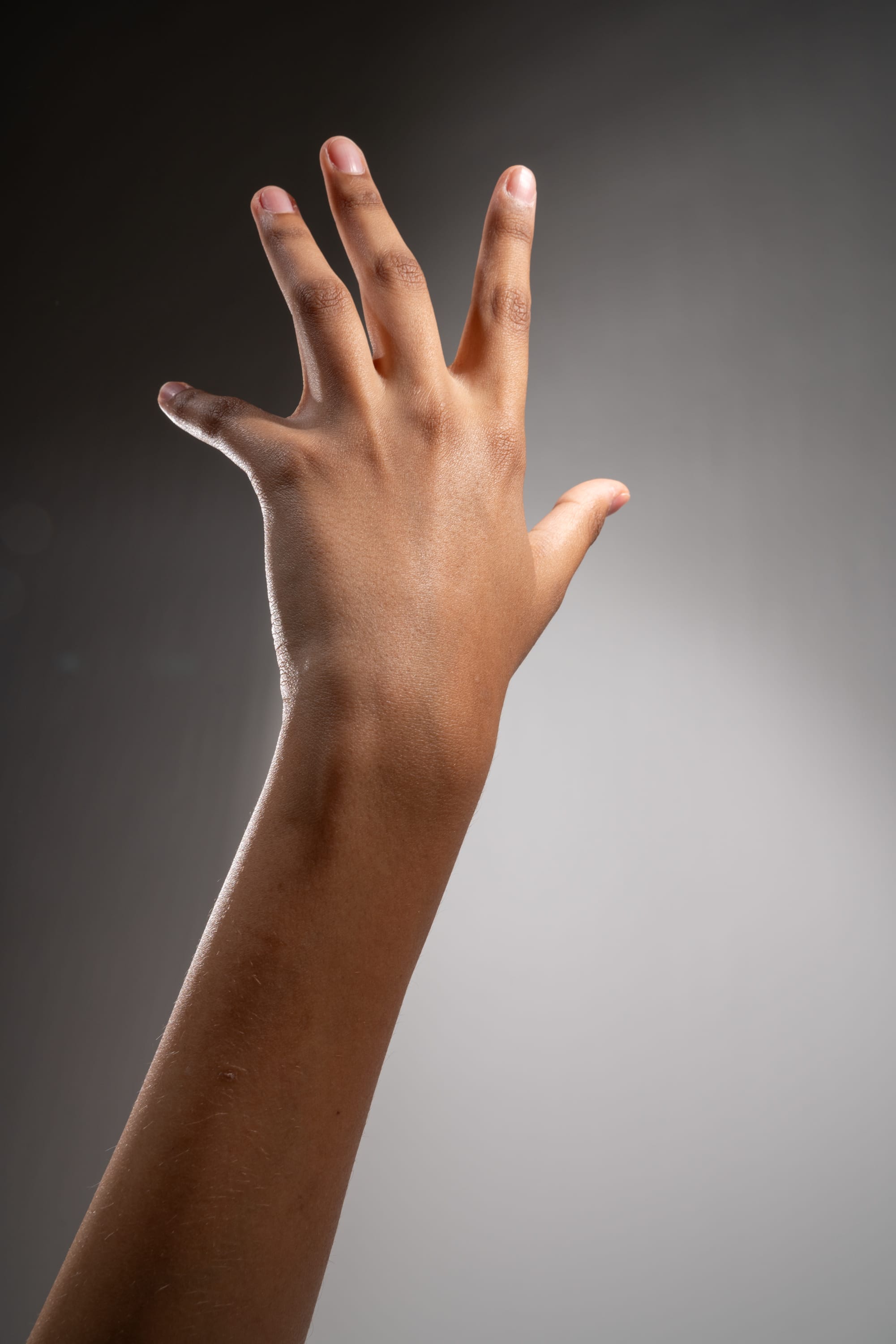 a moderately darkskinned person's left arm and hand are reaching up in front of a gray background that is slightly darker in the upper portion and lighter in the lower portion of the image. the fingers are extended, and the hand is open. the lighting comes from the upper left of the scene.