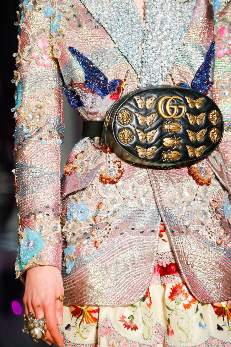 loseup detail shot of a model wearing a highly embellished pink and iridescent jacket and skirt.  the jacket is heavily adorned with beading, embroidery, and sequins featuring floral motifs and bird designs.  the model is also wearing a gucci belt bag/fanny pack, which is black leather with gold lion heads, butterfly, and insect accents. the bag is prominently displayed against the intricately decorated clothing. the overall aesthetic is luxurious, ornate, and highly detailed, suggestive of high fashion runway styling.
