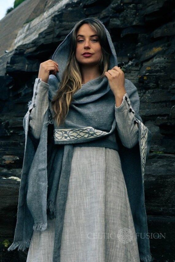 a woman with fair hair is wearing a gray hooded cloak with a decorated belt over a long gray dress in front of a dark, rocky background.