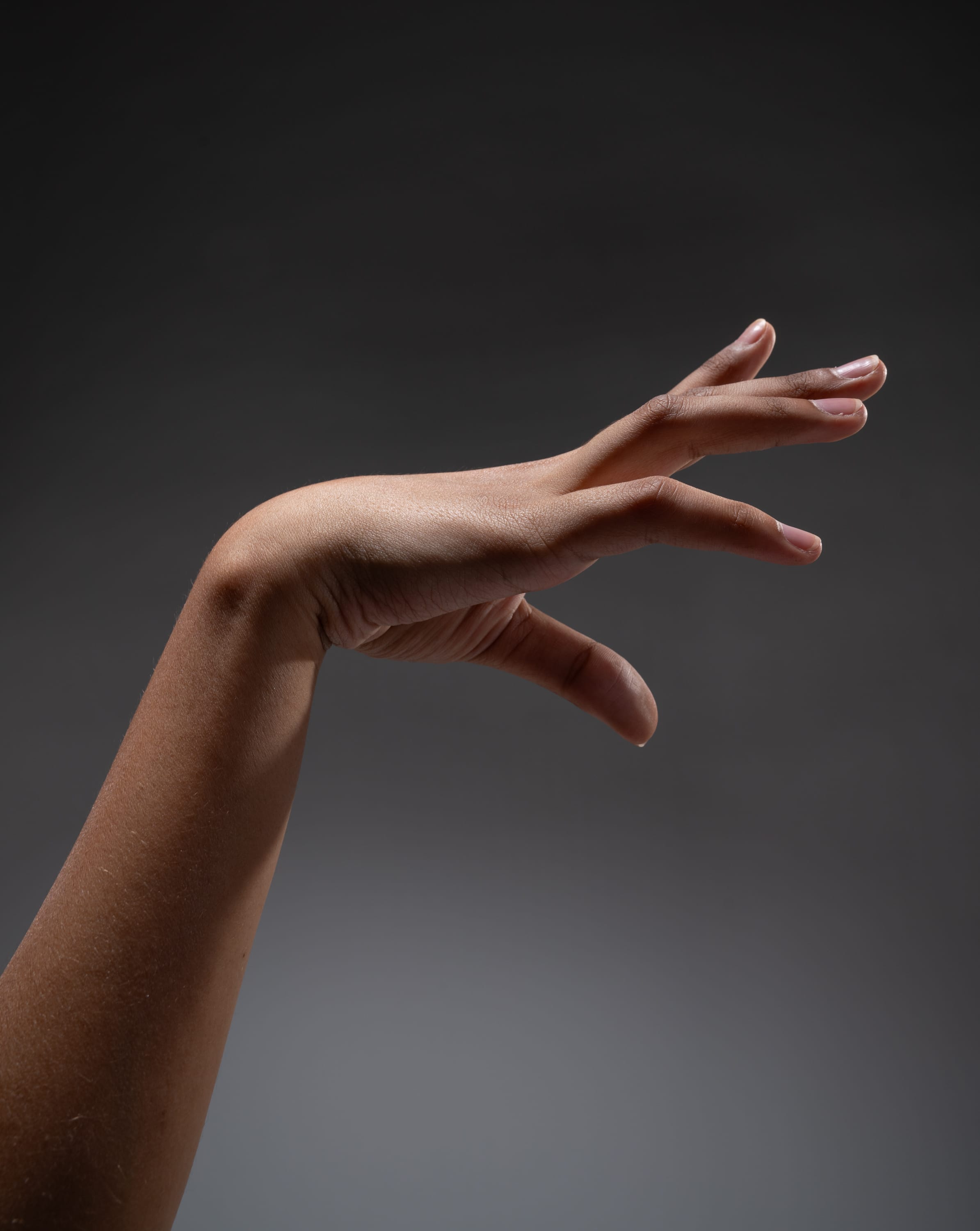 the image showcases a single human arm, bent at the elbow, with the hand elegantly outstretched. the hand is positioned with fingers slightly splayed, creating a sense of delicate reach or expression. the arm, with its dark skin tone, is illuminated by soft studio lighting that highlights the contours and texture of the skin.  the background is a smooth, dark gray, providing a neutral backdrop that emphasizes the arm as the central focus.