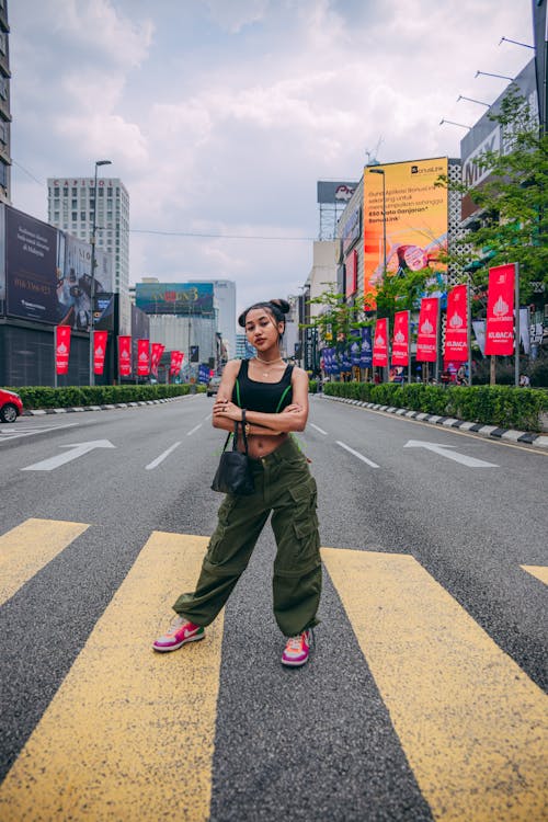 a young asian woman in stylish cargo pants and sneakers stands confidently in a city crosswalk. she is posing with her arms crossed, wearing a black top and carrying a small handbag.  the background features modern city buildings, billboards, and cloudy skies.  the overall mood is urban and fashionable.