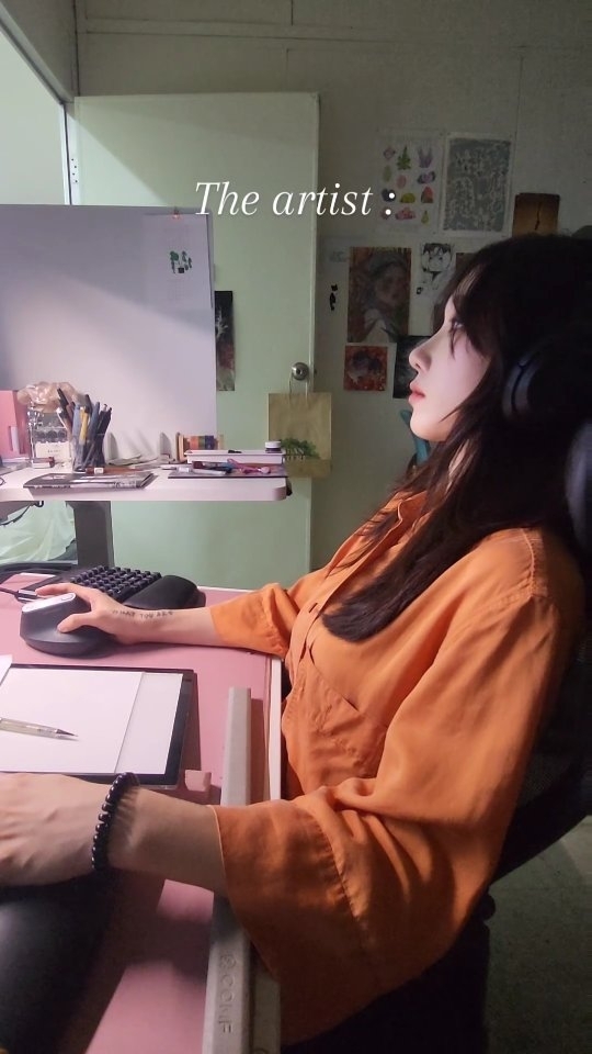 the image shows a young woman sitting at her desk, working on a digital art project. she's wearing an orange shirt and headphones, and appears to be focused on her work.  her workspace is tidy but personalized, with art supplies and other items visible. the text "the artist :" is superimposed on the image, indicating the subject's creative profession. the overall mood is calm and productive.