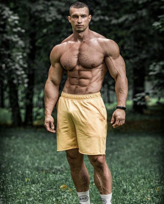 a muscular man in yellow shorts stands in a park, looking directly at the camera. he has a sculpted physique and is posing with his hands on his hips.