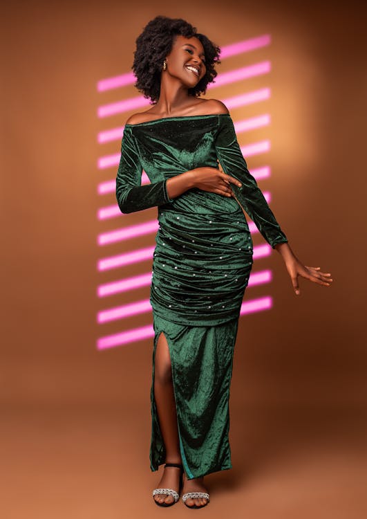 a full shot captures a smiling black woman wearing a green velvet offtheshoulder dress with long sleeves and a slit. she has short, curly hair and is wearing heels and earrings. the background is brown, and there is pink neon light behind her.
