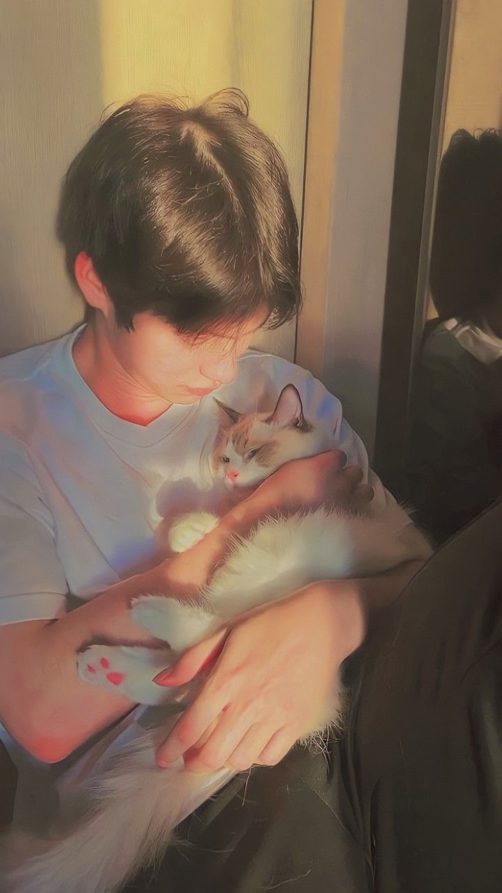 the image captures a heartwarming moment between a young man and his white ragdoll cat. bathed in the warm, golden glow of what appears to be late afternoon sun, the man gently cradles his feline companion. the cat, with its striking blue eyes and soft fur, seems utterly content in the man's arms, emphasizing the special bond they share. the photo has a soft, almost vintage quality, enhancing the overall sense of comfort and love.