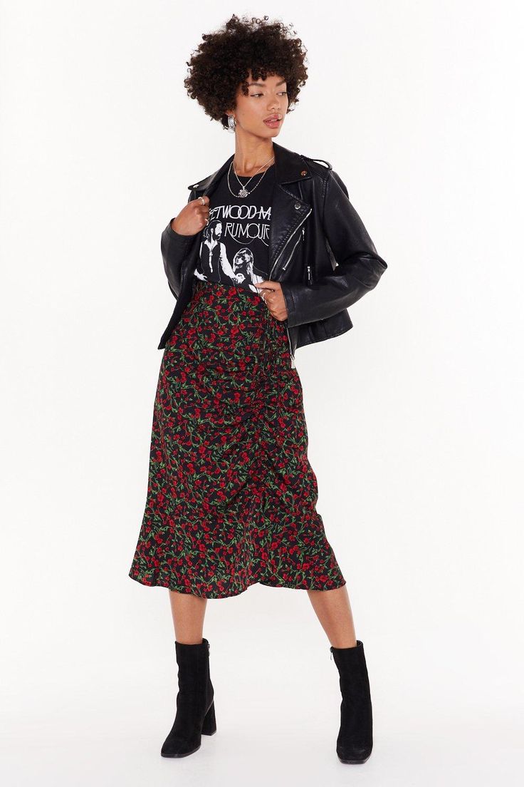 a young brunette woman is posing for the camera. she is wearing a black leather jacket, a black and white graphic tshirt, a floral midi skirt and black ankle boots. she has a bright smile on her face and her hair is styled in a slightly messy wave.