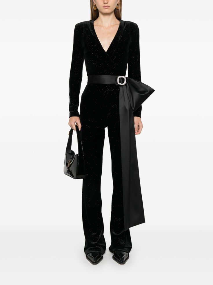 a woman in a black glitter velvet jumpsuit with a matching black bow belt. she is wearing black high heels and holding a black handbag. the model is posing in a studio, with a white background.