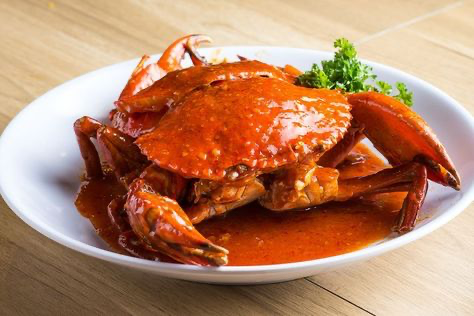 a closeup shot showcases a delectable chilli crab dish, a culinary specialty of singaporean cuisine.  the succulent crab is generously coated in a vibrant, rich red chili sauce, glistening with oil.  the sauce appears thick and flavorful, hinting at a spicy and savory taste.  the crab sits on a white plate, which is positioned on a light brown wooden table. a small garnish of parsley adds a touch of freshness. the overall presentation is enticing and highlights the dish's vibrant colors and textures. the lighting and focus are sharp, creating a visually appealing image perfect for showcasing the dish to potential diners.
