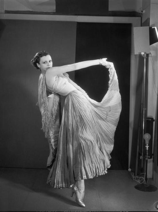 a black and white photograph of a woman in a pleated, flowing gown, possibly a dancer, posing in a studio setting. the dress is lightcolored and billows around her as she holds one arm up. the background features a dark backdrop and visible lighting equipment. the overall aesthetic suggests a vintage photograph from the 1930s. the woman's posture and dress convey elegance and grace.