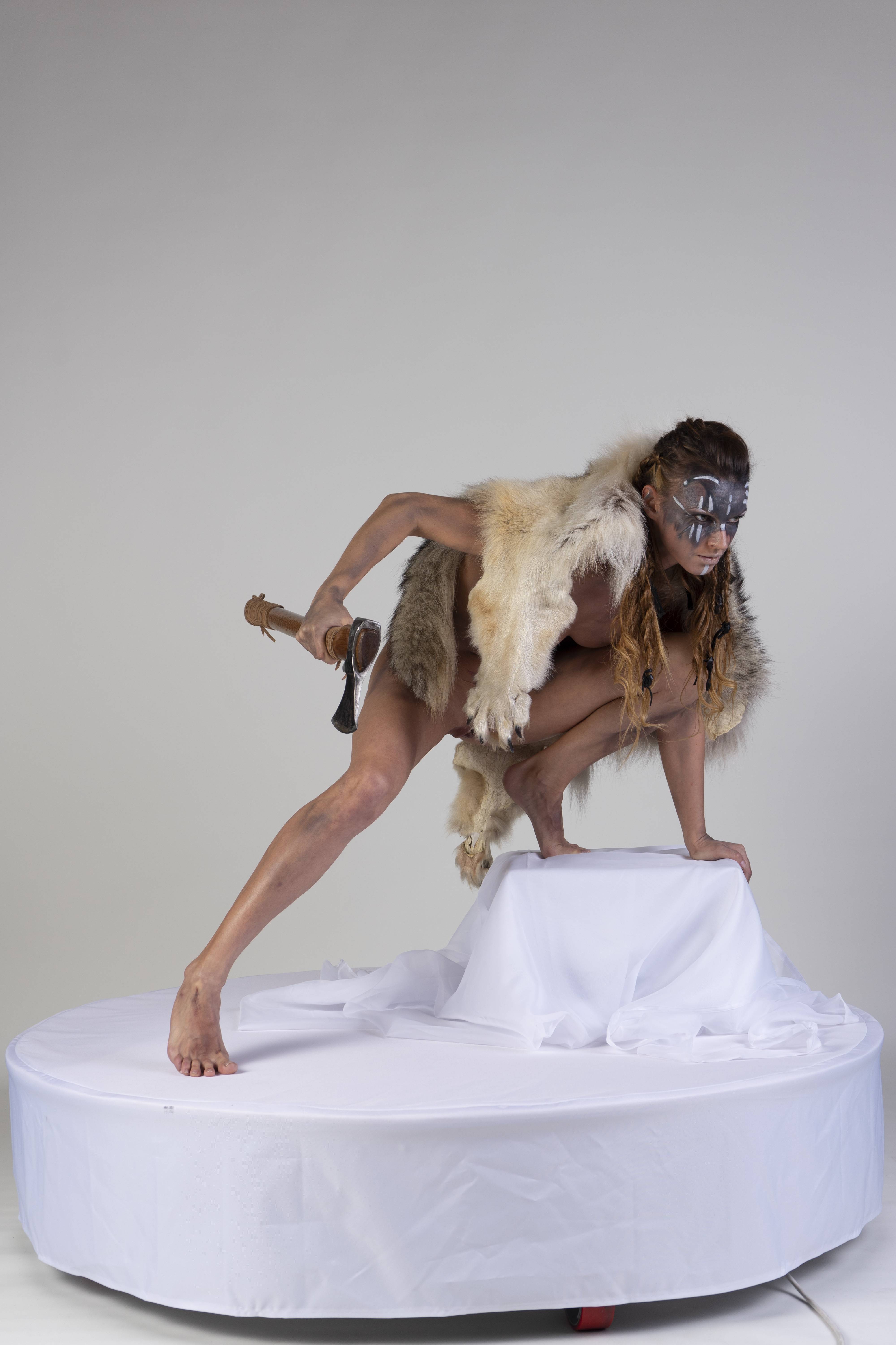 a woman dressed in a primitive warrior costume poses on a platform against a white background. she is barefoot and wearing fur and body paint. she is holding an axe and crouching.